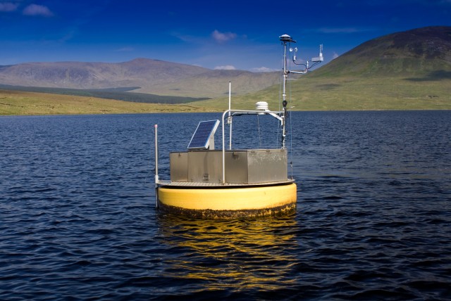 Monitoring buoy with solar panel and sensors floating on a lake surrounded by mountains.