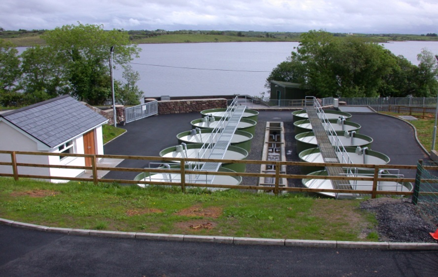 Outdoor aquaculture facility with rows of circular tanks and walkways beside a lake.