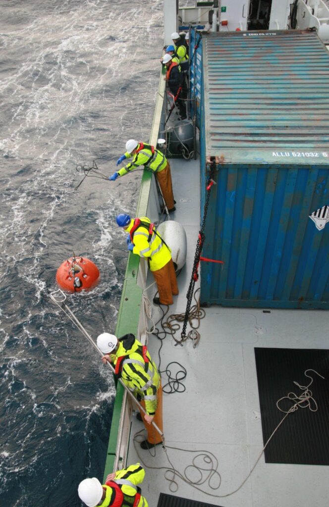 Crew in high-visibility clothing handle lines beside an orange ocean-monitoring instrument floating alongside a research vessel.