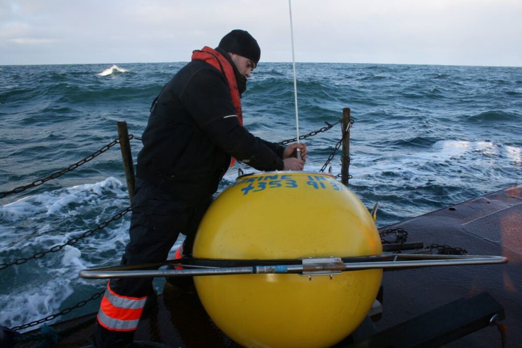 Crew member securing equipment to a large yellow wave buoy on the deck of a vessel in rough seas.