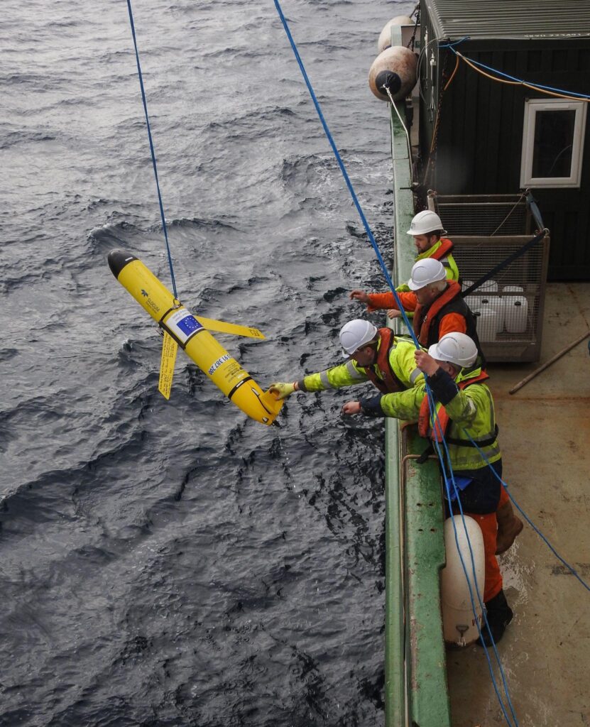 Crew members lower a yellow autonomous underwater glider from the side of a vessel into rough seas.
