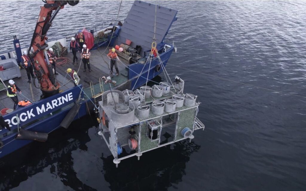 Crew members use a ship-mounted crane to lower a large metal frame of SmartBay monitoring equipment into the sea.