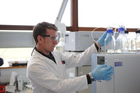 Laboratory analyst wearing safety glasses and gloves operates testing equipment.