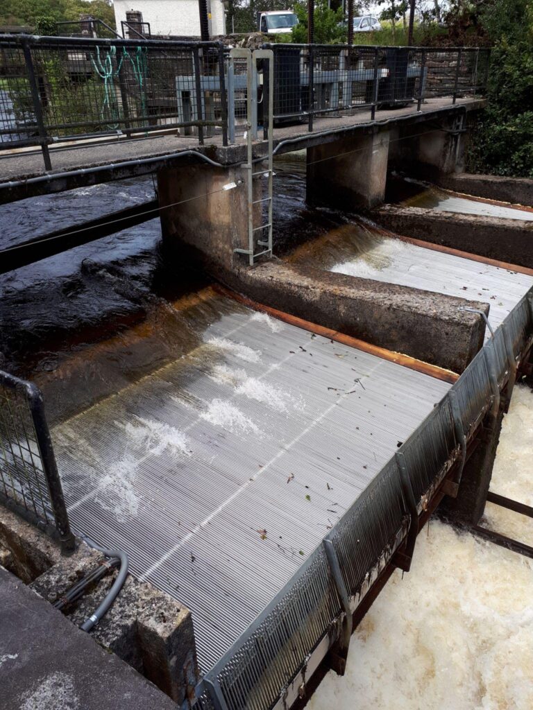 Metal-screened fish passage channels carrying fast-flowing water beneath a fenced monitoring platform.