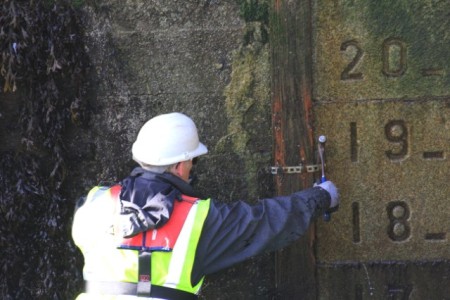 Technician in a hard hat and high-visibility vest checks equipment beside a numbered water-level scale on a harbour wall.