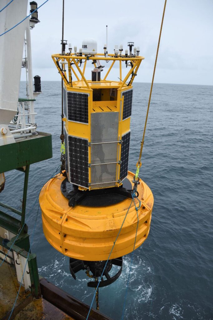 Yellow marine data buoy suspended beside a vessel, with solar panels and monitoring instruments mounted above its float.