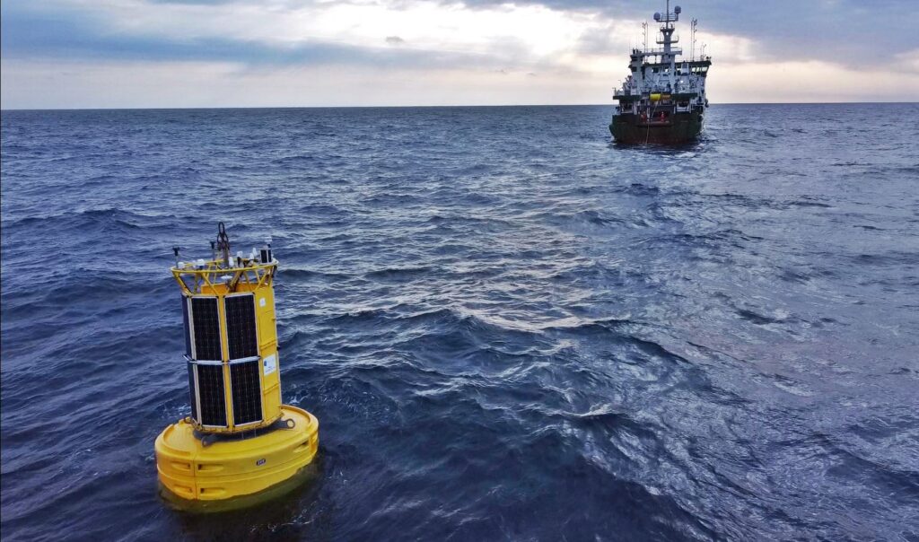 Yellow solar-powered data buoy floating in open water, with a marine research vessel in the distance.