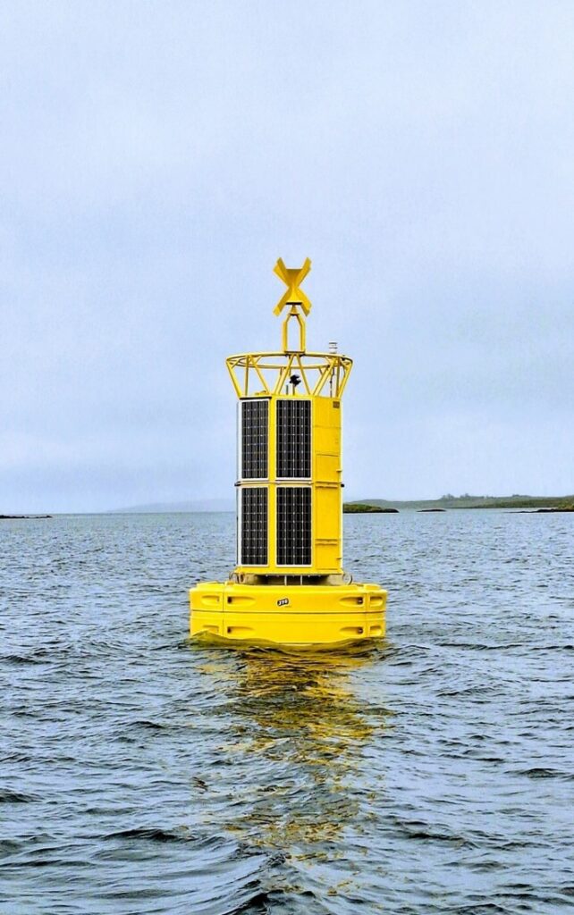 Yellow solar-powered ocean monitoring buoy floating near a low-lying coast.